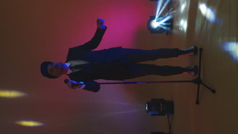 Vertical-video.-Stylish-Man-in-a-jacket-singer-with-a-microphone-stand-dancing-and-singing-in-the-neon-light-in-the-Studio.-An-energetic-and-expressive-singer